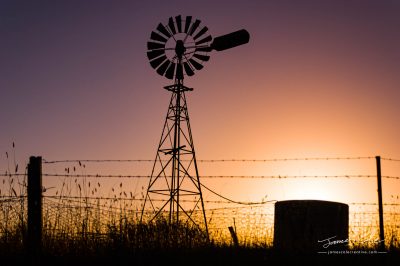 JCCI-100602 - Classic silhouette Australian windmill on farmland behind barbed wire fence at sunset.