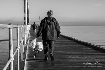 Fisherman walking along a wooden pier on a calm sea with fishing rods and bucket in Frankston Victoria Australia Black and white